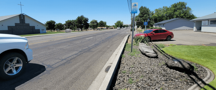 Image Description: A bus stop along a wide arterial suburban road that is placed in gravel next to a curb. There is no sidewalk, ramp, or curb cut, so the stop is completely inaccessible.