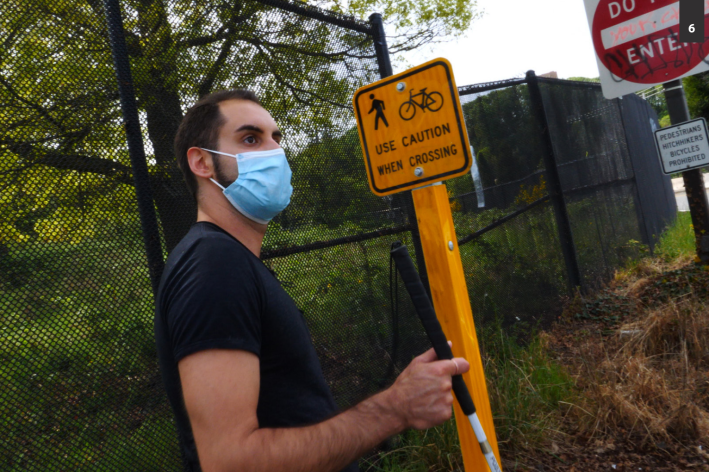 : A brown-skinned man standing at a highway offramp holding a white cane,next to a small yellow traffic sign with an icon of pedestrian crossing and a bicycle reading “Use Caution When Crossing.” Behind that is a large red and white sign reading Do Not Enter and below that a small white sign with black text reading “Pedestrians Hitchhikers Bicycles Prohibited.”