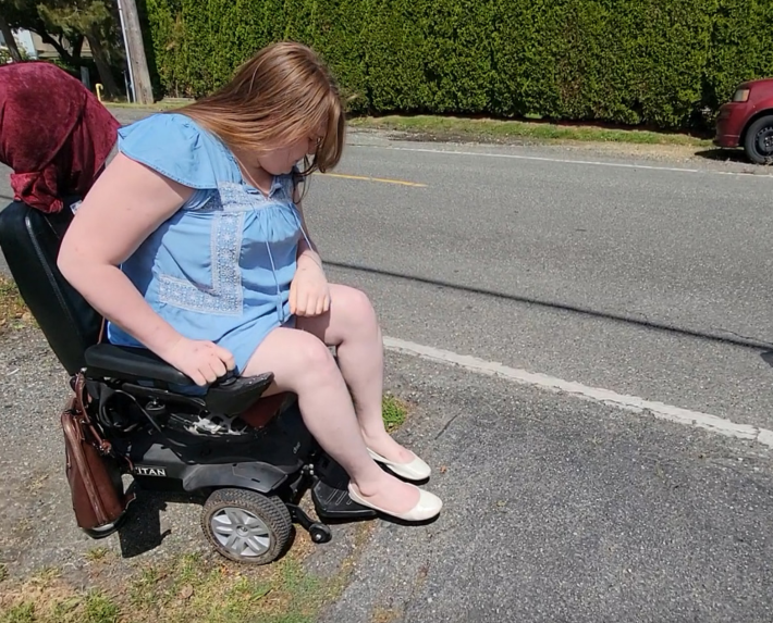 Image description: A light-skinned woman in apowerchair looking at the gravel her chair is stuck in on the side of a road that has no sidewalk.