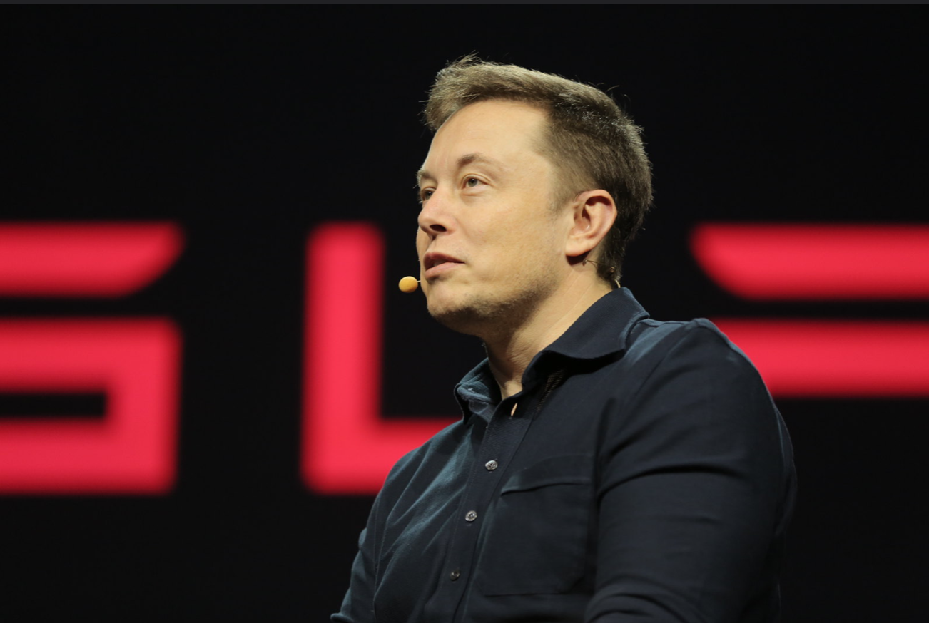 Elon Musk sits on a stage in front of a Tesla logo.