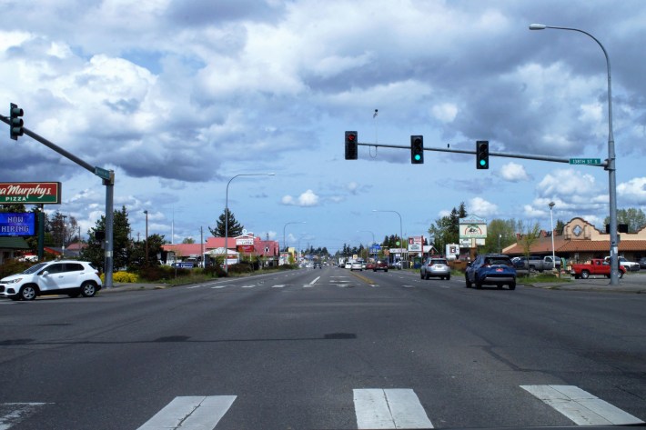 A wide, unwalkable arterial road, lined with strip malls. Several large cars drive along it.