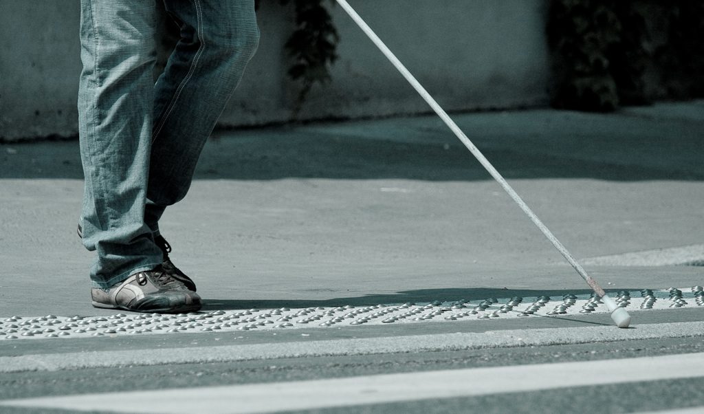 A blind person with a white cane approaches a crosswalk. We can see them from the knee down only.